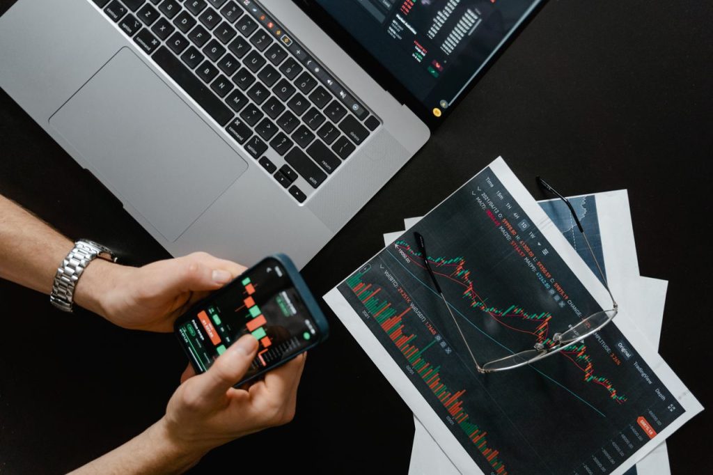 Mastering the First Impression: Your intriguing post title goes here Hands using smartphone beside laptop with stock charts, showcasing digital trading.